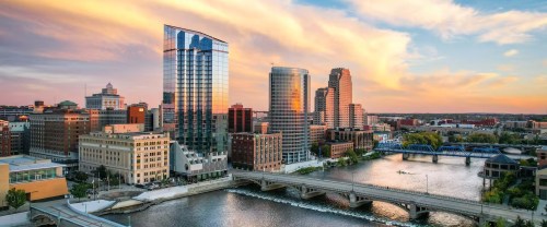 A vibrant city skyline at sunset, featuring modern buildings and a river with a bridge in the foreground.