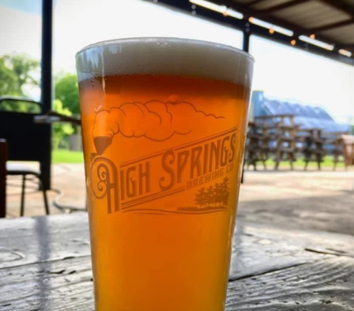 A glass of amber beer with a frothy head, featuring the logo of High Springs Brewing Co. in a rustic setting.