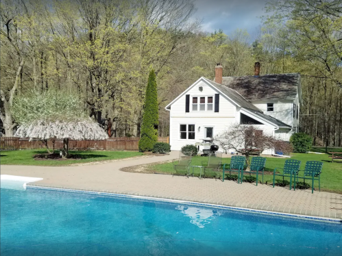 A serene backyard with a pool, a house, and trees, featuring a flowering tree and green chairs by the water.