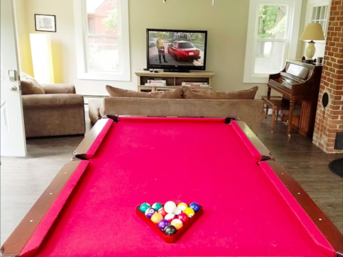A red pool table in a cozy living room with a TV showing a scene and a piano in the background.