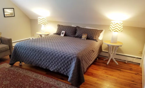 Cozy bedroom with a gray quilted bed, two lamps, a chair, and a decorative rug on wooden flooring.
