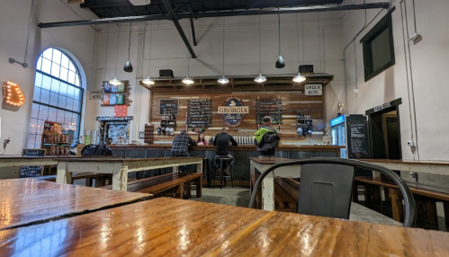 Interior of a brewery with a wooden bar, patrons seated, and a menu board on the wall. Warm, inviting atmosphere.