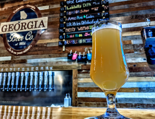 A glass of beer sits on a wooden bar, with a chalkboard menu of drinks in the background at Georgia Beer Co.