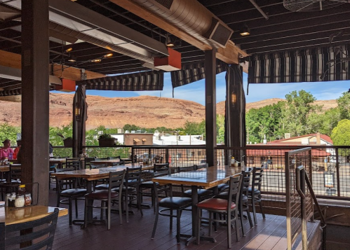 Outdoor dining area with wooden tables and chairs, overlooking a scenic view of hills and buildings.
