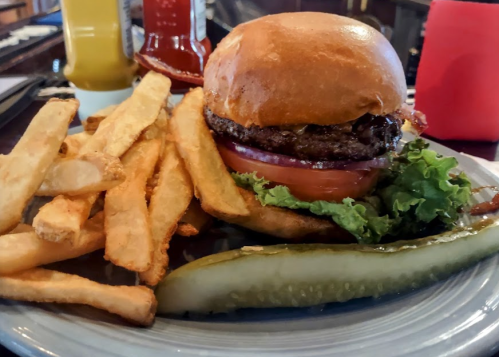 A juicy burger with lettuce, tomato, and pickles, served with golden fries on a plate. Condiments in the background.