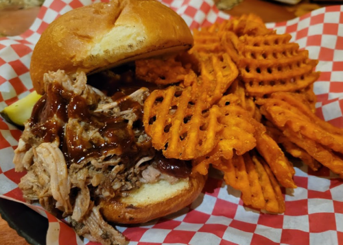 A pulled pork sandwich with barbecue sauce on a bun, served with a side of crispy waffle fries in a red-checkered basket.