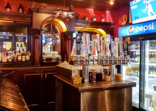 A bar with a wooden counter, various beer taps, shelves of liquor, and a Pepsi fridge in a cozy setting.