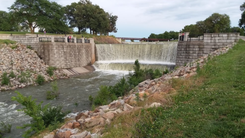A waterfall cascades over a stone dam, surrounded by greenery and rocky banks under a cloudy sky.