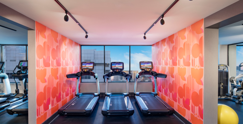A modern gym with three treadmills against a colorful wall and large windows showing a city view.