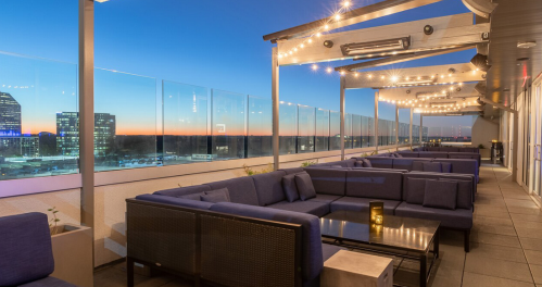 Rooftop lounge with blue seating, city skyline, and sunset view, illuminated by string lights.