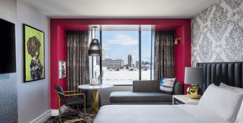 Modern hotel room featuring a pink accent wall, large window, stylish furniture, and city views.