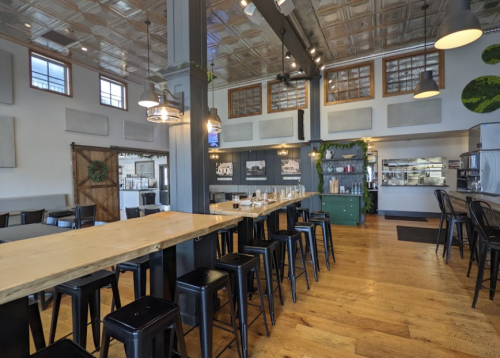 A spacious, modern restaurant interior featuring a long wooden bar, metal stools, and a bright, open layout.