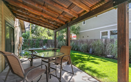 A shaded patio with a table and chairs, surrounded by greenery and a well-maintained lawn.
