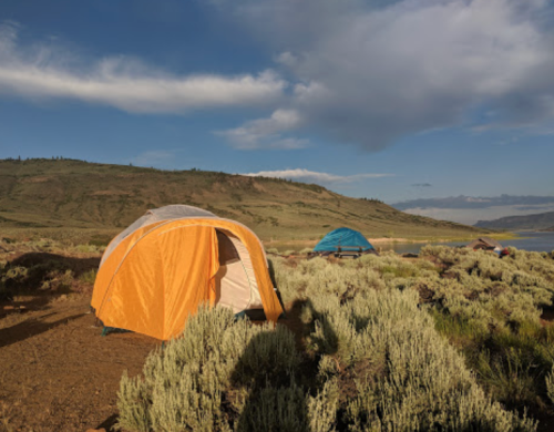 Two tents set up in a grassy area near a lake, with hills and a blue sky in the background.