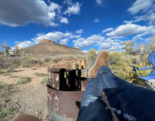 A person relaxes with feet up on a rusted barrel, overlooking a mountain under a blue sky with fluffy clouds.