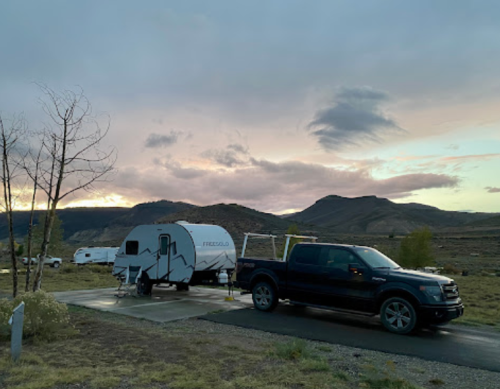 A camper trailer parked beside a pickup truck, set against a backdrop of mountains and a cloudy sky at dusk.