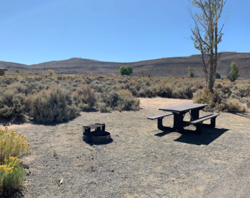 A picnic table near a fire pit in a dry, open landscape with sparse vegetation and distant hills under a clear blue sky.