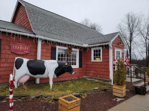 A red barn-style building with a cow statue out front, decorated for the holidays, and surrounded by greenery.