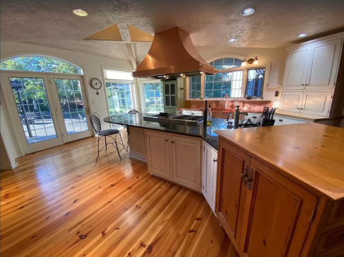 Bright kitchen with wooden floors, large windows, a central island, and a stylish range hood. Outdoor view visible.