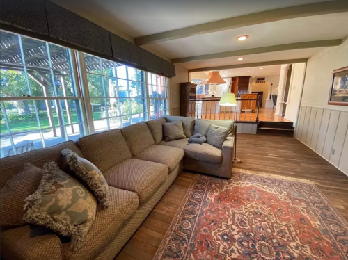 Cozy living room with a sectional sofa, lamp, and a view of a bright outdoor space through large windows.