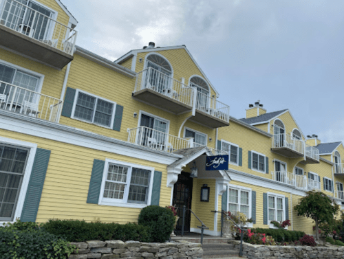 A yellow hotel building with balconies, white trim, and a welcoming entrance surrounded by greenery and flowers.