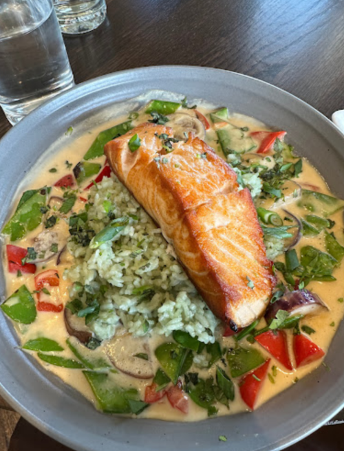 A plate of salmon on a bed of green rice, surrounded by colorful vegetables in a creamy sauce.