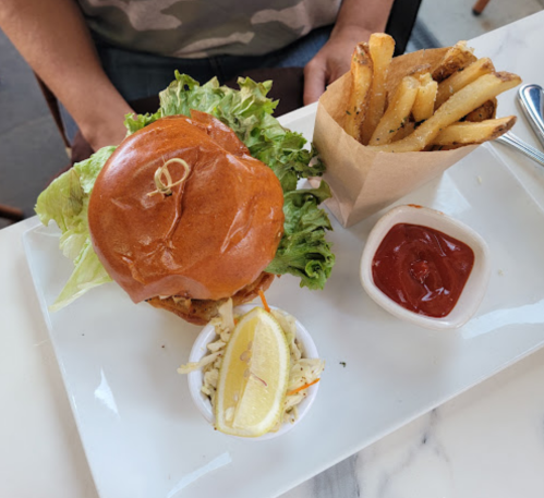 A burger with lettuce on a plate, accompanied by fries in a paper cup, coleslaw, and a lemon wedge.