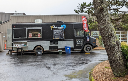 A black food truck parked in a lot, with a menu displayed and trees nearby, on a rainy day.