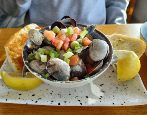 A bowl of clams topped with tomatoes and green onions, served with lemon wedges and garlic bread on the side.