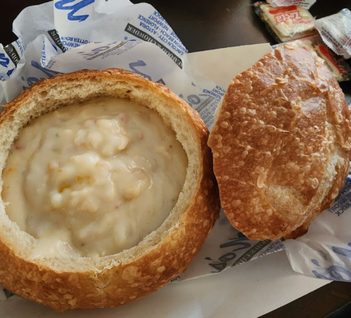 A bread bowl filled with creamy soup, accompanied by the lid of the bread, on a paper wrapper.