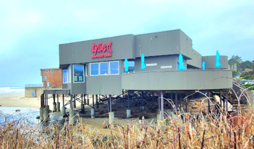 A seafood grill on stilts by the beach, with a cloudy sky and ocean waves in the background.
