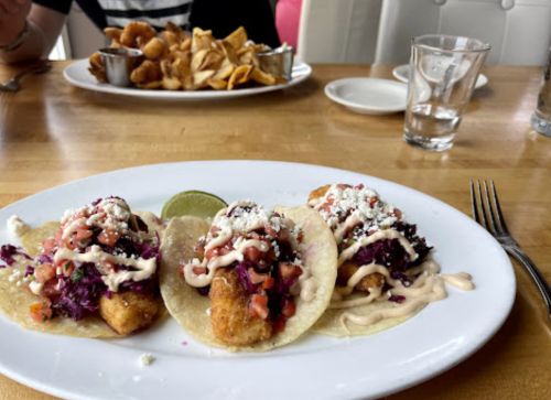 Three fish tacos topped with cabbage, pico de gallo, and sauce, served with a lime wedge on a white plate.