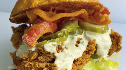 Close-up of a crispy fried chicken sandwich with bacon, pickles, lettuce, and creamy sauce on a toasted bun.