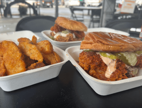 Two chicken sandwiches with pickles and sauce, alongside a serving of crispy potato wedges in white containers.
