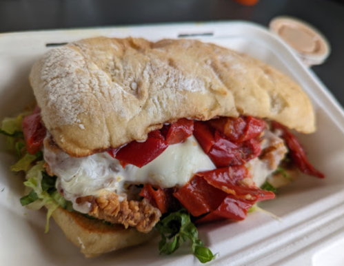 A sandwich with fried chicken, lettuce, roasted red peppers, and a fried egg, served in a soft bread roll.