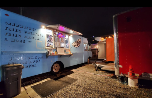 A blue food truck at night, with an "Open" sign, serving sandwiches, fried potatoes, and fried chicken.