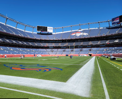 A wide view of a football stadium with blue seats, a green field, and the Denver Broncos logo on the turf.