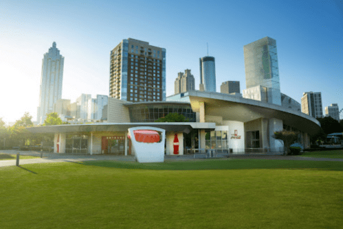 Modern building with a curved design, surrounded by green grass and city skyscrapers in the background, under a clear blue sky.