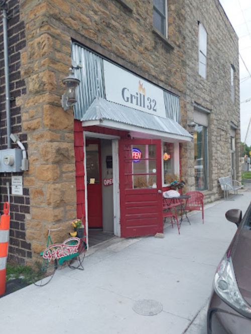 A rustic restaurant entrance with a red door, sign reading "Grill 32," and outdoor seating on a stone building.