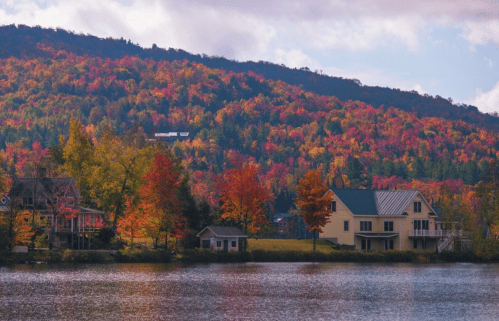 A serene lake scene with colorful autumn trees and charming houses along the shore, set against a vibrant hillside.