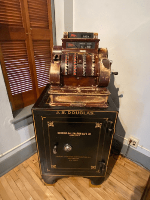 An antique cash register atop a vintage black safe, both showing signs of wear, in a room with wooden flooring.