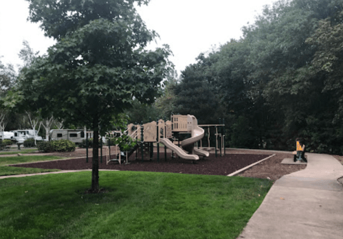 A playground with slides and climbing structures surrounded by grass and trees, with a path leading nearby.