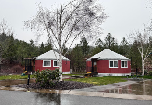 Two red yurts with white roofs in a rainy setting, surrounded by trees and a grassy area.