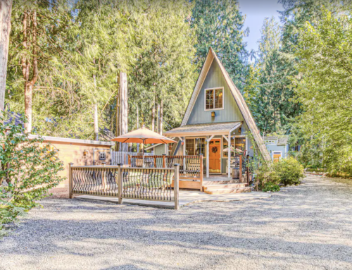 A charming A-frame cabin surrounded by tall trees, featuring a wooden deck and an umbrella for outdoor seating.