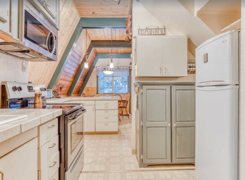 Cozy kitchen with wooden beams, white cabinets, and a dining area visible in a cabin-style setting.