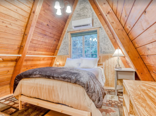 Cozy A-frame bedroom with wooden walls, a large bed, and a window overlooking nature. Warm lighting and rustic decor.