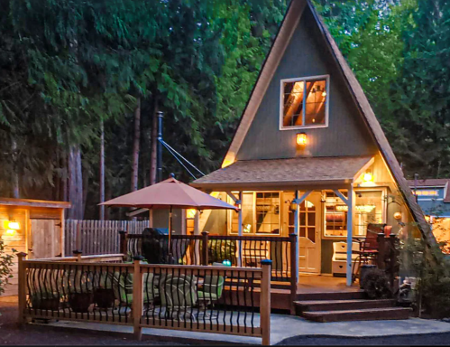 A cozy A-frame cabin surrounded by trees, featuring a deck with seating and warm outdoor lighting at dusk.