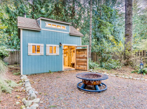 A cozy blue cabin surrounded by trees, featuring a fire pit in front and a wooden door open to the interior.