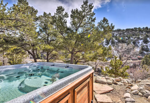 A hot tub surrounded by trees and rocky terrain, with a scenic view of mountains and a blue sky.