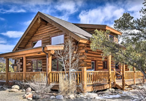 A rustic log cabin with large windows, surrounded by trees and rocky terrain under a blue sky.